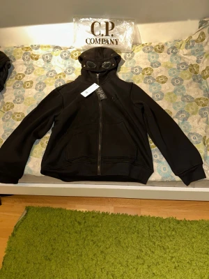 Svart hoodie  från C.P. Company - Svart hoodie från C.P. Company med dragkedja framtill och ikoniska glasögon i huvan. Helt ny. Med tags på. Pris går att diskuteras. Öppen för frågor och bud. Mått kan skickas