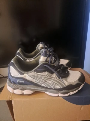Asics gel nyc vita och marinblå sneakers - Snygga sneakers från Asics i vitt och marinblått med silverdetaljer. Skorna har en klassisk låg siluett, meshpaneler för bra ventilation och robust sula för extra komfort. Perfekta för dig som gillar sportig stil och vill ha något bekvämt till vardags.