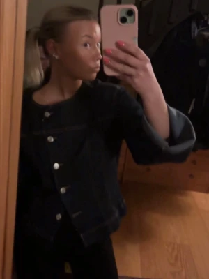 Mörkblå jeansjacka med silverknappar - Snygg mörkblå jeansjacka med raka ärmar och stora silverfärgade knappar framtill. Klassisk modell utan krage som ger en modern look. Perfekt att slänga över en t-shirt eller hoodie för en avslappnad stil.