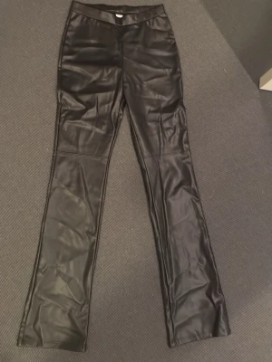 Svarta fuskskinnbyxor från Lindex - Snygga svarta byxor i fuskskinn från Lindex. Byxorna har raka ben och resår i midjan för skön passform. Perfekta för dig som vill ha en trendig och edgy look. Materialet är glansigt och ger en cool vibe till outfiten.