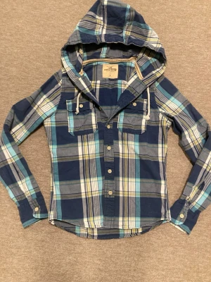  Hollister Hoodie - Snygg blårutig skjorta från Hollister med huva och knappar framtill. Skjortan har två bröstfickor med lock och långa ärmar. Tillverkad i mjuk bomull, perfekt för en avslappnad och cool stil.