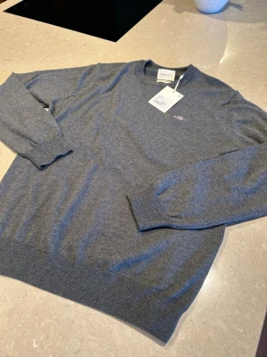 Grå stickad tröja från Gant - Snygg grå stickad tröja från Gant med rund halsringning och diskret logga broderad på bröstet. Tröjan har långa ärmar, ribbade muddar och nederkant. Perfekt basplagg i mjukt material som passar till det mesta.