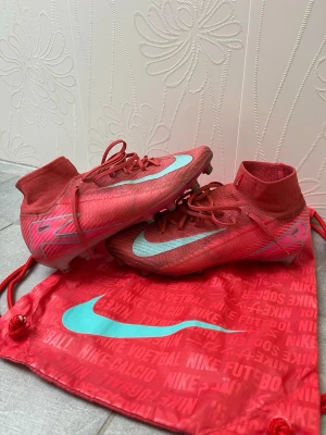 Röda Nike Mercurial Elite fotbollsskor  AG - Säljer ett par röda Nike Mercurial elite AG fotbollsskor med turkosa detaljer och högt skaft. Skorna har snörning och är tillverkade i syntetmaterial för bra grepp och känsla på planen. Medföljer matchande röd Nike-påse med turkos logga. Köptes för 3000kr. Storlek 42.5