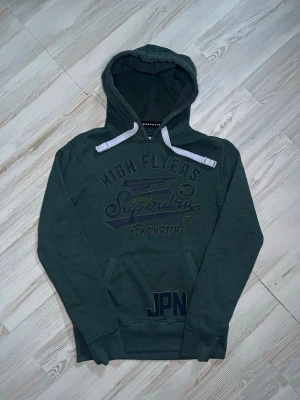 Grön hoodie från Superdry - Cool grön hoodie från Superdry med tryck 'High Flyers' och 'Yokohama' på bröstet🤩Längd 70cm bredd 53cm  Hoodien är i fint skick och säljs för ett bra pris🙌🏻 Hör av er vid funderingar samt snabbaffär😇