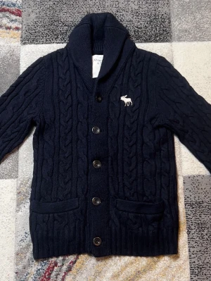 Mörkblå kabelstickad kofta Abercrombie - Mörkblå kabelstickad kofta från Abercrombie & Fitch med sjalkrage, stora knappar och två fickor framtill. Vit broderad logga på bröstet. Tjockt och mjukt material som håller värmen, perfekt för kyliga dagar.