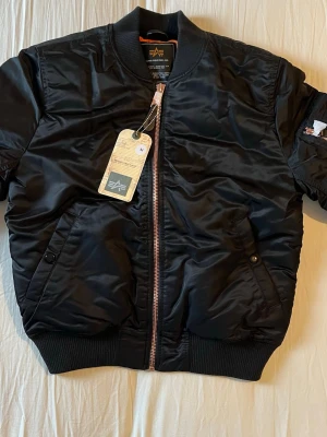 Svart bomberjacka från Alpha Industries - Stilren svart bomberjacka från Alpha Industries med klassisk orange innerfoder, ribbade muddar och dragkedja i koppar. Jackan är helt oanvänd och kommer med påse och tags. Nypris: 2999kr, Mitt pris: 1499kr 🤝