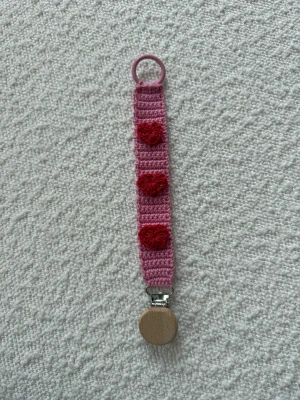 Rosa virkad napphållare med hjärtan - Söt handvirkad napphållare i rosa med tre röda hjärtan som dekoration. Hållaren har en rund träklämma och en plastögla för enkel fastsättning. Perfekt accessoar för att hålla koll på nappen och ge en gullig touch till looken.