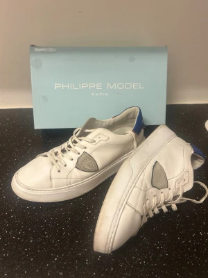 Vita Philippe Model sneakers - Snygga vita sneakers från Philippe Model med klassisk låg silhuett. Skorna har gråa detaljer, blå hälflik och ikonisk sköldlogga på sidan. Tillverkade i läder med vita snören och diskret design som passar till det mesta.
