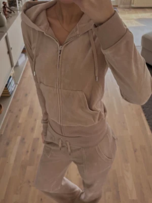 Beige velourset från Juicy Couture - Mysigt beige velourset från Juicy Couture med dragkedja, huva och fickor på tröjan samt matchande byxor med dragsko i midjan. Perfekt för chill dagar hemma.  Storlek S i byxorna och M i koftan . Använd enstaka gånger 