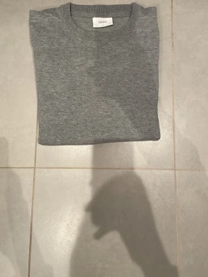 Grå tröja från Gant rundhalsad - En enkel och snygg grå tröja från Gant med rund halsringning och ribbade muddar. Tröjan har en klassisk rak passform och är tillverkad i ett mjukt stickat material som känns skönt mot huden. Perfekt som basplagg till olika stilar.