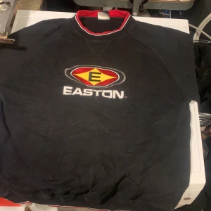 Svart Easton sweatshirt med logga - Cool svart sweatshirt från Easton med stor broderad logga i gult, rött och vitt på bröstet. Tröjan har rund hals med röd och vit kant samt ribbade muddar. Perfekt för dig som gillar sportig stil och vill sticka ut lite extra.