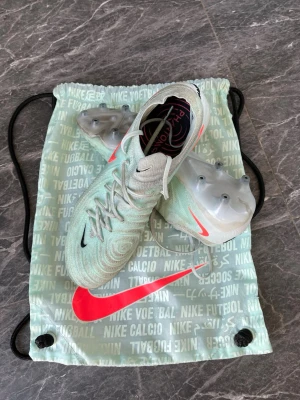 Nike Phantom GX mint fotbollsskor Elite  - Snygga Nike Phantom GX fotbollsskor i mintgrönt med neonrosa swoosh och detaljer. Skorna har en modern, strömlinjeformad design och är tillverkade i syntetmaterial för bästa bollkontroll. Sulan har fasta dobbar för spel på naturgräs. Perfekta för dig som vill sticka ut på planen.