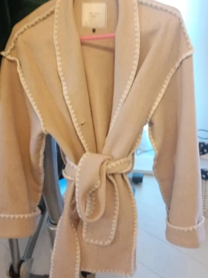 Beige kappa med knytskärp Neo Noir - Stilren beige kappa från Neo Noir med markerade sömmar i vitt och ett brett knytskärp i midjan. Kappan har bred krage och trekvartsärmar, vilket ger en avslappnad men trendig look. Perfekt för dig som gillar minimalistisk och modern stil.