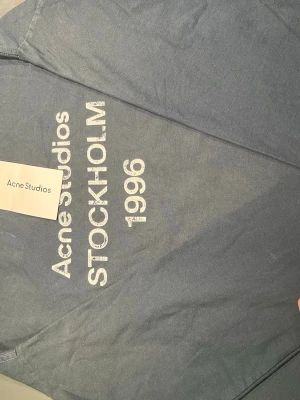 Blå Acne Studios t-shirt med tryck - Snygg blå t-shirt från Acne Studios med vitt tryck framtill där det står 'Acne Studios STOCKHOLM 1996'. Klassisk rund halsringning och korta ärmar. Perfekt för dig som gillar stilrena plagg med en urban touch.