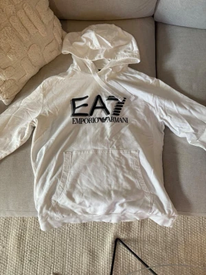 Vit hoodie från Emporio Armani EA7 - Snygg vit hoodie från Emporio Armani EA7 med stor logga framtill. Klassisk huva med dragsko och magficka. Tillverkad i mjukt material som känns skönt mot huden. Perfekt för en avslappnad och trendig look.