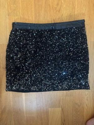 Svart paljettkjol från Mango - Glittrig svart kortkjol från Mango täckt med paljetter. Kjolen har en rak passform och bred linning i satinliknande material. Perfekt för dig som vill sticka ut och glänsa lite extra.