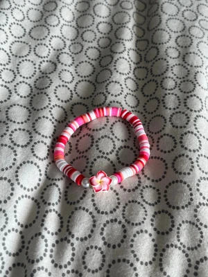 Färgglatt pärlarmband med blomma - Sött armband med platta pärlor i rosa, vitt och rött samt två silverfärgade pärlor. I mitten sitter en detalj i form av en blomma i vitt, rosa och gult. Armbandet är elastiskt och passar de flesta handleder. Perfekt för dig som gillar färg och unika accessoarer.