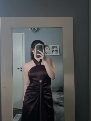 Mörkbrun långklänning med halterneck - Elegant mörkbrun långklänning i satinliknande material med halterneck och dekorativ detalj i midjan. Klänningen har en åtsittande passform upptill och ett vackert fall. Perfekt för speciella tillfällen.