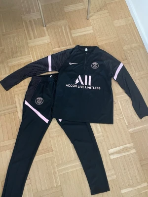 Svart PSG träningsset från Nike - Säljer ett svart träningsset från PSG och Nike med rosa detaljer. Setet består av långärmad tröja med halv dragkedja och matchande byxor. PSG-logga och Nike swoosh på både tröja och byxor. Materialet är troligtvis polyester, perfekt för träning eller chill.