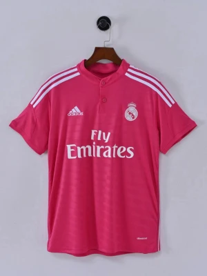 Rosa Real Madrid tröja Adidas - Snygg rosa Real Madrid matchtröja från Adidas med vita detaljer och tryck. Tröjan har korta ärmar, rund hals med knapp och klassiska Adidas-ränder på axlarna. Materialet är lätt och andas, perfekt för träning eller match. Fly Emirates-logga på bröstet.