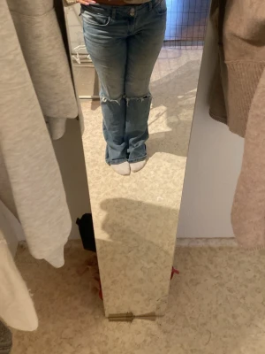 Blå bootcut jeans med slitningar - Säljer ett par blå bootcut jeans med slitningar vid knäna och klassisk femficksdesign. Jeansen har låg midja och är tillverkade i denim med en lätt tvättad look. Perfekta för dig som gillar en avslappnad och trendig stil.