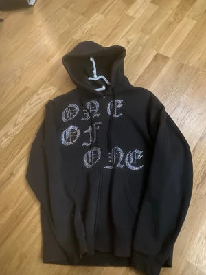 Svart hoodie med strasstext - Säljer en svart hoodie med dragkedja och huva. På framsidan står det 'ONE OF ONE' i stora bokstäver dekorerade med strass. Hoodien har en avslappnad passform och fickor framtill. Perfekt för dig som gillar streetwear och vill sticka ut.