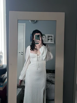 Vit långklänning med puffärmar - Superfin vit långklänning med puffiga långärmar, snörning framtill och slits. Klänningen har en romantisk känsla med diskret prickigt mönster och markerad midja. Perfekt för sommarens tillfällen.