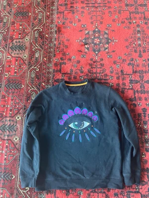 KENZO sweatshirt med ögonmotiv - Cool svart sweatshirt med ett stort ögonmotiv i blått, vitt och lila på bröstet. Tröjan har rund halsringning och långa ärmar. Materialet ser ut att vara mjuk bomull eller bomullsblandning. Perfekt för dig som gillar unika prints och streetwear vibes. Finns tyvärr ingen tagg i tröja  eftersom den var jobbig för nacken under användning 