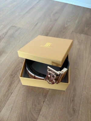Randigt Burberry bälte med guldspänne 110cm - Säljer ett klassiskt Burberry bälte med det ikoniska randiga mönstret i beige, svart, vitt och rött. Bältet har ett stort guldigt 'TB'-spänne och är gjort i skinn. Perfekt accessoar för att lyfta vilken outfit som helst.