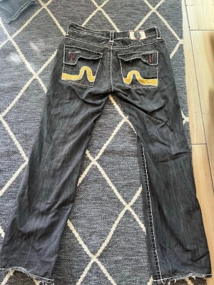 Svarta Nudie Jeans med gula detaljer - Säljer ett par svarta Nudie Jeans med raka ben och tydliga gula broderier på bakfickorna. Jeansen har kontrastsömmar och ficklock med röda knapphål. Klassisk denimkänsla och cool streetstil.