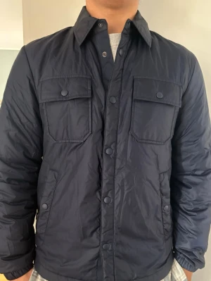 Fodrad jacka från woolrich  - Stilren mörkblå vindjacka med två bröstfickor med lock och knappar samt sidofickor. Jackan har klassisk krage, lång ärm och stängs med knappar framtill. Perfekt till hösten och i använd endast ett fåtal gånger. Nytt skick