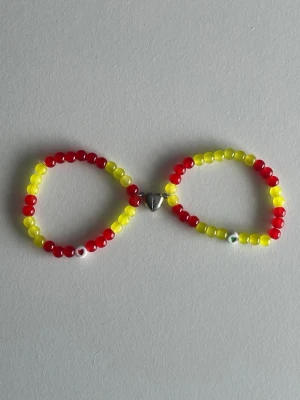 Gulgrönt och rött pärlarmband med magnet-hjärta - Säljer ett handgjort armband med glada pärlor i rött och gult. Armbandet har ett unikt silverfärgat hjärtlås i mitten och små detaljer som ögonpärlor. Perfekt för dig som gillar färg och vill sticka ut med dina accessoarer.