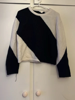 Svartvit stickad tröja med diagonal design - Säljer en stickad tröja i svart och vitt med diagonal färgblock-design. Tröjan har rund halsringning, långa ärmar och en avslappnad passform. Perfekt för dig som gillar grafiska mönster och vill sticka ut lite extra.