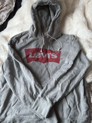 Grå hoodie från Levi's med logga - Säljer en grå hoodie från Levi's med den klassiska röda loggan på bröstet. Hoodien har huva med dragsko och en stor magficka. Tillverkad i mjukt bomullsmaterial, perfekt för en avslappnad och cool stil.