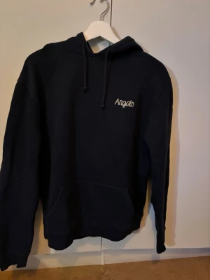 Mörk blå hoodie från Axel Arigato - Svart hoodie från Axel Arigato med broderad logga framtill och en stor färgglad brodyr på ryggen som föreställer ett landskap med träd och gul äng. Hoodien har huva med dragsko och en stor magficka. Perfekt för dig som vill ha en stilren men unik look.