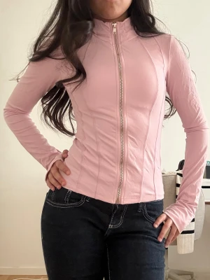 Ljusrosa figurnära ziptröja - Supersnygg ljusrosa ziptröja med figurnära passform och långa ärmar. Tröjan har en hel dragkedja framtill och markerade sömmar som ger en sportig look. Perfekt att styla med jeans eller kjol för en trendig outfit.