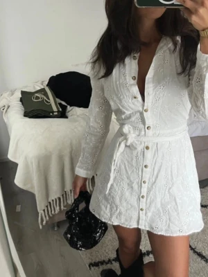 Vit  klänning med broderi och knappar - Säljer en vit klänning med broderade blommor, knappar framtill och knytskärp i midjan. Klänningen har lång ärm och v-ringning, perfekt för en fräsch och somrig look. Passar dig som gillar feminina detaljer och stilren design.