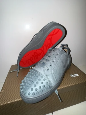 Gråa Christian Louboutin sneakers - Snygga gråa sneakers från Christian Louboutin med ikoniska röda sulor och coola nitar på tån. Skorna är i mocka och har matchande grå skosnören. Perfekta för dig som vill sticka ut med en edgy och lyxig stil.