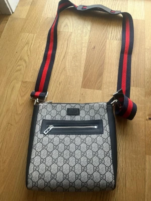 Gucci axelväska med GG-mönster - Snygg axelväska från Gucci med klassiskt GG-monogram i grått och svart. Väskan har en justerbar axelrem i rött och marinblått, silverfärgade detaljer och en praktisk ytterficka med dragkedja. Perfekt för dig som vill ha en trendig och lyxig accessoar.