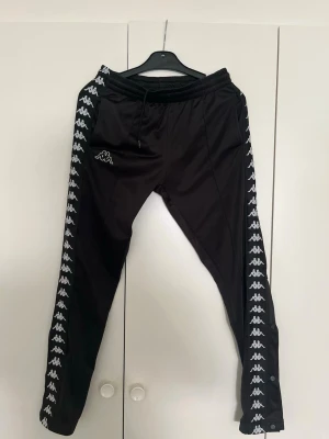 Svarta Kappa mjukisbyxor med logga - Säljer ett par svarta mjukisbyxor från Kappa med vita loggor längs båda benen och dragsko i midjan. Byxorna har en sportig look och är tillverkade i ett glansigt, mjukt material. Perfekta för chill eller streetwear-stil.