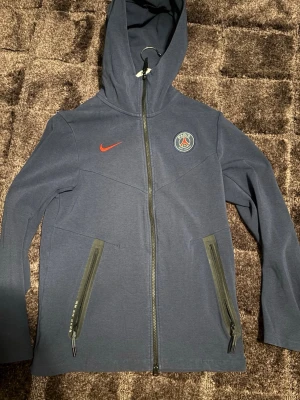 Mörkblå PSG hoodie från Nike - Snygg mörkblå hoodie från Nike med PSG-logga på bröstet och röd Nike-swoosh. Dragkedja framtill och två fickor med dragkedja. Tillverkad i mjukt material och har en huva med dragsko. Perfekt för dig som gillar sportig stil och Paris Saint-Germain.