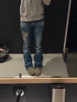 Blåa lågmidjade jeans med tryck - Säljer mina favorit jeans. blåa lågmidjade jeans med coolt tryck i vitt och guld på vänster lår.  låg midja och slitningar. Perfekta för dig som gillar streetstyle och vill sticka ut. Säljer för att de har blivit försmå. Köpte för 900 spänn