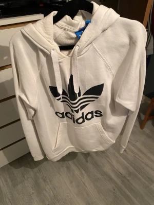 Vit Adidas hoodie med stor logga - Säljer en vit hoodie från Adidas med stor svart Trefoil-logga och text på bröstet. Hoodien har huva med dragsko och en klassisk känguruficka framtill. Tillverkad i mjukt material som känns skönt mot huden. Perfekt för en avslappnad streetwear-look.