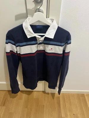 Randig långärmad skjorta från Gant - Mörkblå långärmad skjorta från Gant med vita, röda och blå ränder över bröstet. Skjortan har vit krage och knäppning framtill. Perfekt för en sportig och avslappnad stil.