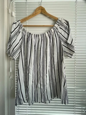 Randig offshoulder blus i vitt och svart - Säljer en snygg offshoulder blus med svarta vertikala ränder på vit bakgrund. Blusen har korta ärmar som är skurna och gör axeln bar,  och en lös passform med resår runt axlarna. Perfekt för en trendig och avslappnad look. Materialet känns lätt och luftigt.