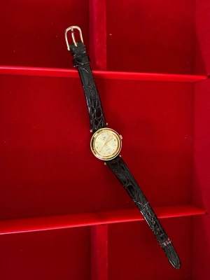 Guldig quartzklocka med svart läderband - Stilren klocka med rund guldfärgad boett och ljus urtavla dekorerad med små stenar som timmarkeringar. Svart band i mönstrat läder ger en klassisk look. Perfekt accessoar för dig som gillar tidlös design.