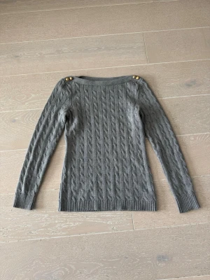 Grå kabelstickad tröja Lauren Ralph Lauren - Snygg grå kabelstickad tröja från Lauren Ralph Lauren med guldfärgade knappar på axlarna. Tröjan har lång ärm, ribbade muddar och en klassisk, smal passform. Perfekt för dig som gillar stilrena och tidlösa plagg.