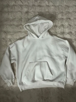 Vit hoodie med magficka - Klassisk vit hoodie med stor huva och rymlig magficka. Tröjan har ribbade muddar vid ärmslut och nederkant samt lång ärm. Perfekt för en avslappnad och enkel stil.