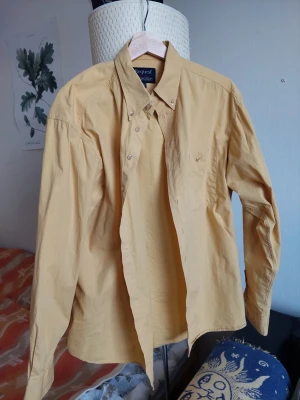 Gul skjorta - Klassisk uppknäppt skjorta med gul färg som ger retro- och sommarkänsla. Äldre modell från ett av kappahls egna märken, hög kvalitet i materialet, som ny. Avslappnad passform, kan bäras utanför byxor eller instoppade.