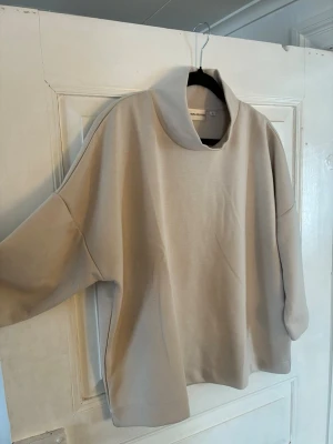 Beige tröja med hög krage Carin Wester - Snygg beige tröja från Carin Wester i storlek M. Tröjan har en avslappnad passform, hög krage och  halva ärmar. Tillverkad i mjukt material som känns skönt mot huden. Perfekt för dig som gillar minimalistisk och clean stil.